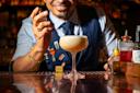 A bartender in a vest and tie sprays garnish over a frothy cocktail in a coupe glass, with various bar tools and bottles on the counter. The scene is warmly lit, capturing a joyful expression.