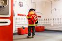 A young child dressed as a firefighter stands in a playroom with fire department gear and a wall decorated with simple line drawings of buildings and trees.