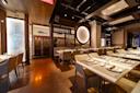 A modern restaurant dining area with neatly set tables and beige chairs, warm lighting, wood accents, wall art featuring a fish, and a wine display in the background. The atmosphere is elegant and inviting.