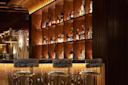 A stylish bar with a backlit wooden shelf displaying assorted liquor bottles, three patterned cushioned bar stools, and warm ambient lighting creating a cozy atmosphere.