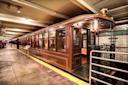 A vintage subway train from 1907 is displayed in a well-lit station with tiled walls. The wooden exterior and classic design evoke a historical feel. A metal railing and information plaque are also visible.