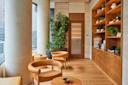 A modern, serene waiting area with tan circular chairs, small wooden tables, a large potted plant, and a wall with shelves holding towels and small plants. Tall windows with blinds illuminate the space, complemented by a wooden floor.