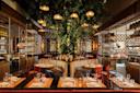 Elegant restaurant interior with wooden tables set for dining, cushioned round booth seating, red chairs, shelves with glassware, and a large tree with hanging lights as a centerpiece. Warm, ambient lighting throughout.