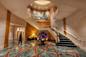 A luxurious hotel lobby featuring a grand spiral staircase with a golden railing. The floor has an intricate geometric design. A large floral arrangement is centered beneath the staircase. A person walks towards the elevators.