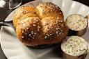 A small skillet of golden brown rolls topped with seeds, served on a plate with two ramekins of creamy dips, garnished with herbs and seeds. The presentation is appetizing and elegant.