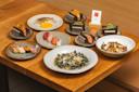 A wooden table set with assorted Japanese dishes, including sushi, nigiri, grilled fish, hand rolls with seaweed, pasta, and a plate of octopus, displayed on ceramic plates.