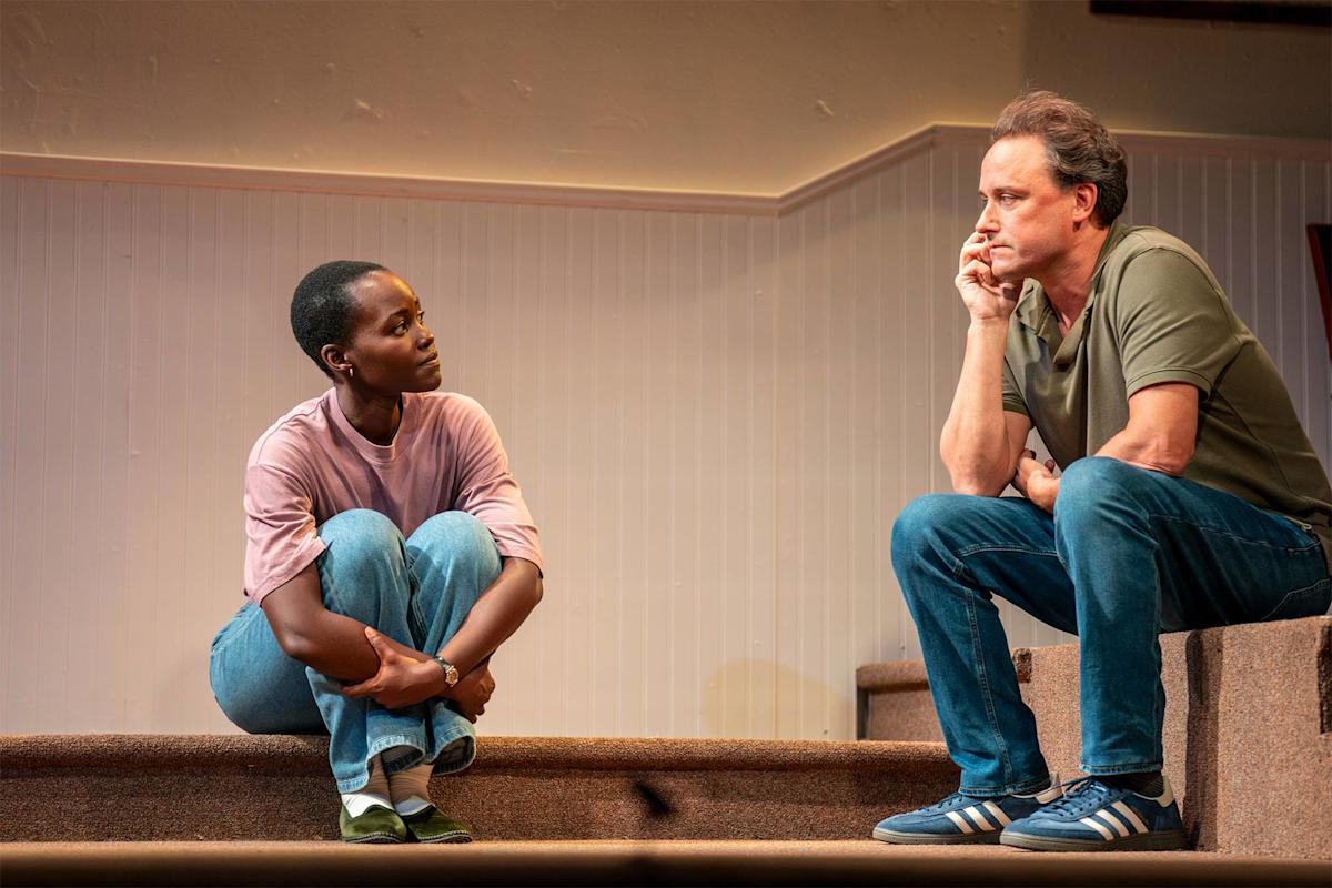 A woman in a pink shirt and jeans sits cross-legged on steps, facing a man in a green shirt and blue jeans who sits on a higher step, resting his chin on his hand. They appear to be having a serious conversation.