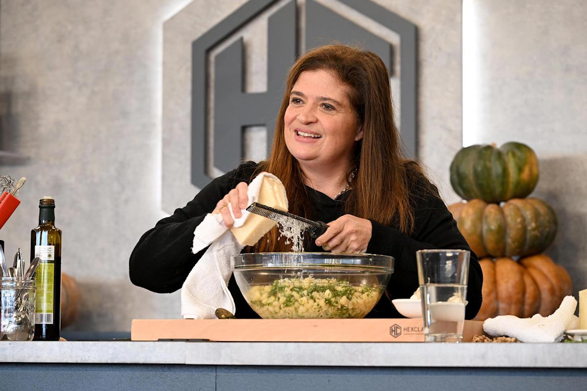 A woman smiles while grating cheese over a glass bowl of food on a kitchen counter. Olive oil, a glass of milk, and pumpkins are visible in the background.