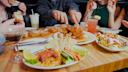 Four people share a meal at a restaurant table, featuring fried chicken, pita with dips and vegetables, salad, assorted drinks, and bread. Hands are seen serving and eating the food.