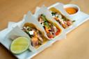 Three tacos filled with meat, vegetables, cheese, and sauce are lined up on a white plate. A lime wedge and a small bowl of orange sauce are on the side, all set on a wooden surface.