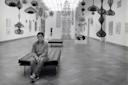 A person sits alone on a bench in a modern art gallery with intricate hanging sculptures and artwork displayed on white walls. The space is bright with a patterned wooden floor and a high ceiling.