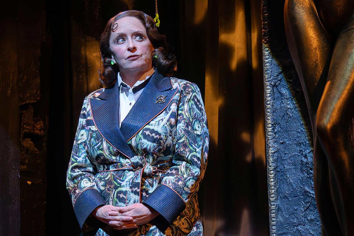 A woman in a patterned suit stands on stage with dramatic lighting, hands clasped, and a thoughtful expression. There is a large sculpture visible to the right and heavy curtains in the background.