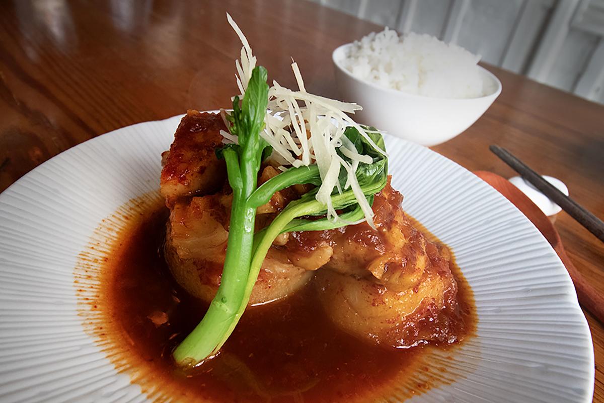 A white plate with braised meat in a rich brown sauce, garnished with green vegetables and shredded root, sits on a wooden table. In the background, there is a small bowl of white rice and a pair of chopsticks.