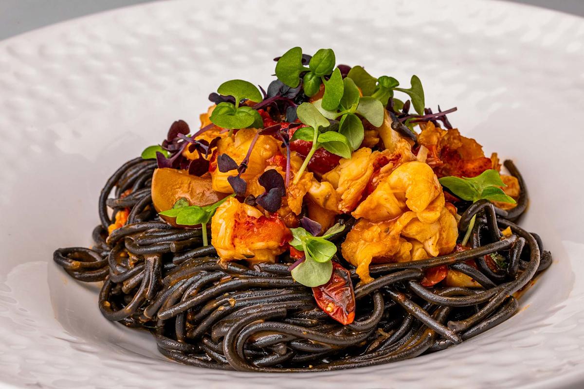 Black squid ink pasta topped with lobster pieces, cherry tomatoes, and microgreens served on a white plate.