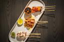 Three skewers with grilled meats and vegetables are arranged on a white oval plate with lime wedges and a small bowl of dipping sauce, all placed on a dark wooden table.