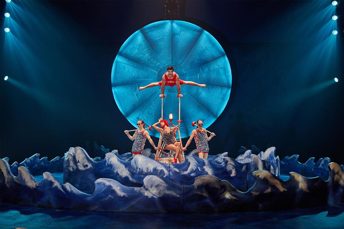 Acrobats perform a balancing act on stage, with one person balancing atop a tall apparatus and four others supporting below. They are dressed in colorful costumes, surrounded by a wave-like set design, under a large, illuminated circular backdrop.