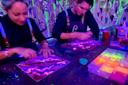Two people sit at a table making neon art on paper under blacklight, surrounded by glowing art supplies and vibrant paint splatters on the wall. Both wear black shirts with aprons and are focused on their artwork.