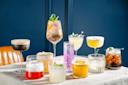 A variety of colorful cocktails displayed on a table, set against a dark blue background. There are ten drinks in different types of glasses, some with garnishes like mint leaves, lemon slices, and ice cubes. Each drink varies in color and presentation.