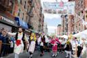 A group of people in traditional Greek attire dance joyfully on a street at a festival. A banner overhead reads "Greek Jewish Festival." Onlookers line the street, enjoying the festive atmosphere