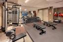 A modern gym with various exercise equipment. The room features weight benches, dumbbells neatly stacked on racks, resistance bands, a cable machine, and stability balls. Mirrors line the back wall, reflecting the neatly organized space.