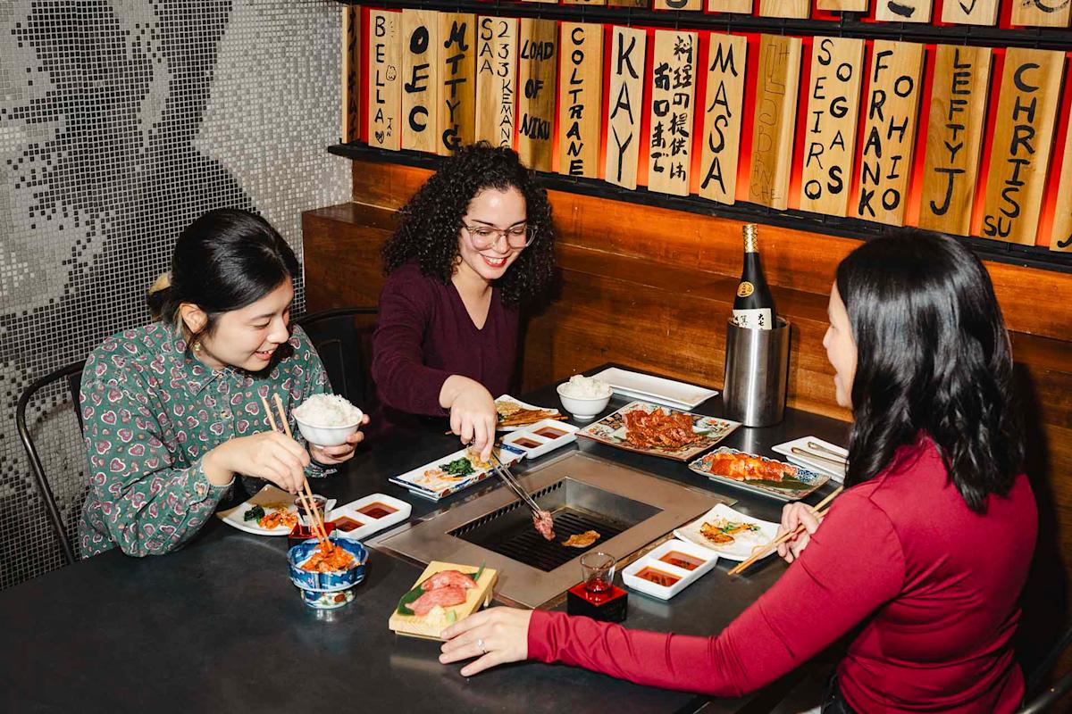 Three people sit around a table cooking and eating food at a Korean BBQ restaurant, with various dishes and sauces on the table and name plaques on the wall behind them.