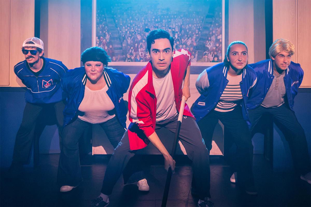 Five people in sports-themed costumes pose in a dramatic, crouched stance on stage, with a background image of a cheering crowd behind them, under bright theatrical lighting.