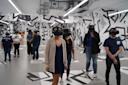 A group of people stands in a modern room with abstract black-and-white wall designs, wearing virtual reality headsets and looking in different directions. Some wear casual clothes; the floor has white rectangular mats.