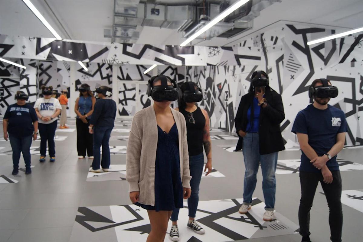 A group of people stands in a modern room with abstract black-and-white wall designs, wearing virtual reality headsets and looking in different directions. Some wear casual clothes; the floor has white rectangular mats.