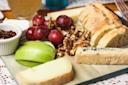 A wooden board with sliced baguette, green apple slices, red grapes, walnuts, assorted cheeses, and a small dish of jam, arranged for a cheese platter.