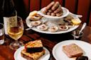 A table set with a glass of white wine, a bottle, grilled bread, pâté, and a platter of oysters on ice topped with sausages, garnished with lemon halves, on a dark wooden surface.