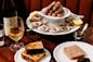 A table set with a glass of white wine, a bottle, grilled bread, pâté, and a platter of oysters on ice topped with sausages, garnished with lemon halves, on a dark wooden surface.