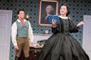 A woman in a black Victorian dress reads from a blue book while a man in period clothing watches her. They are on a stage set resembling a historical room, with a portrait of George Washington on the wall.