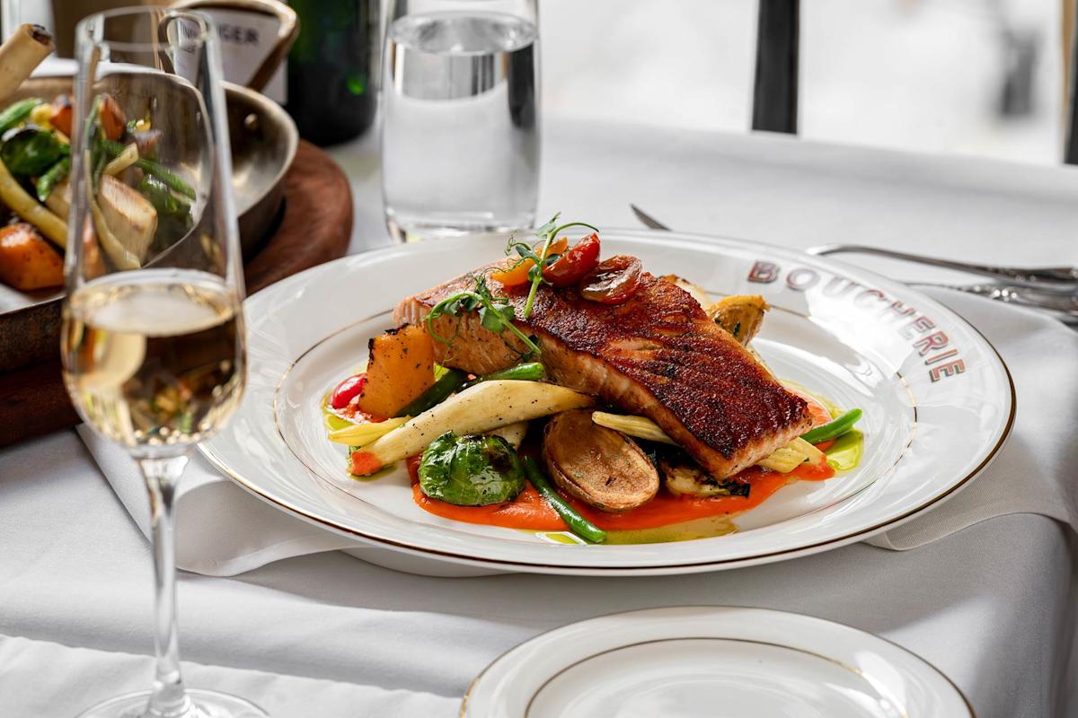 A plate of seared fish with roasted vegetables, garnished with cherry tomatoes and herbs, sits on a white tablecloth next to a glass of white wine and a bowl of mixed vegetables.