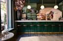 A stylish bar with green paneling, wooden stools, hanging globe lights, and floral arrangements. Bottles and glasses are displayed on shelves behind the counter, and large windows let in natural light.