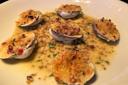 Six baked clams topped with a golden breadcrumb mixture sit on a white plate, surrounded by a mustard and herb sauce with visible mustard seeds and garnished with chopped herbs.