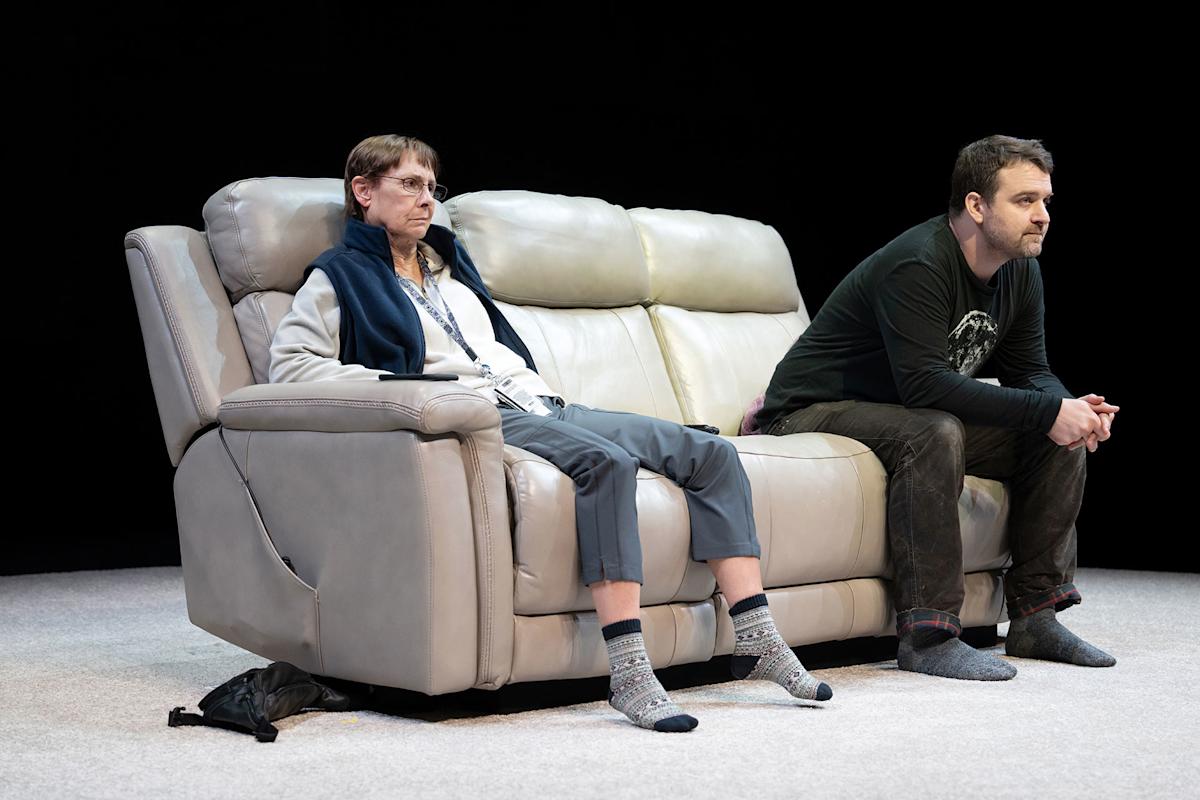 Two people sit on a beige sofa. The person on the left wears glasses, a fleece jacket, and patterned socks, looking tired. The person on the right, in dark clothes, leans forward with hands clasped, both on a light carpeted floor.