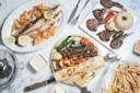 A table set with grilled fish, lamb chops with rice, mixed grilled kebabs with vegetables and flatbread, a plate of fries, lemon slices, and dipping sauces on a marble surface.