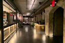 Dimly lit bar interior with exposed brick walls and metal accents. A long bar counter with stools is on the left. Low lighting highlights the modern, industrial design, and a dining area with neatly set tables is visible in the background.