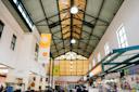Indoor view of Jamaica Market with a high, triangular ceiling, hanging banners, food stalls on the right, and people walking around. Bright natural light comes through large windows at the end.