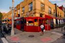 A person walks past a red corner restaurant with outdoor seating on a city street, surrounded by brick buildings, storefronts, and a one-way street sign.