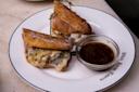 A plate with two halves of a sandwich filled with melted cheese and meat, secured with toothpicks. A small glass bowl of dipping sauce is beside the sandwich, all served on a white plate with "White House Tavern" text.