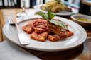 A white plate with rigatoni pasta topped with tomato sauce and fresh basil sits on a wooden table. A fork is placed on the left of the plate. In the background, a bowl with olive oil and another plate of food are partially visible.