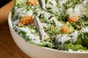 A close-up of a Caesar salad in a white bowl, topped with grated cheese, croutons, anchovies, and chopped herbs on a bed of romaine lettuce. The salad is placed on a wooden surface.