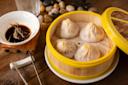 A bamboo steamer holds four steamed dumplings on a wooden surface. Beside it, a small white bowl contains soy sauce with ginger strips. In the background, there are decorative stones and a metal tong rests nearby.