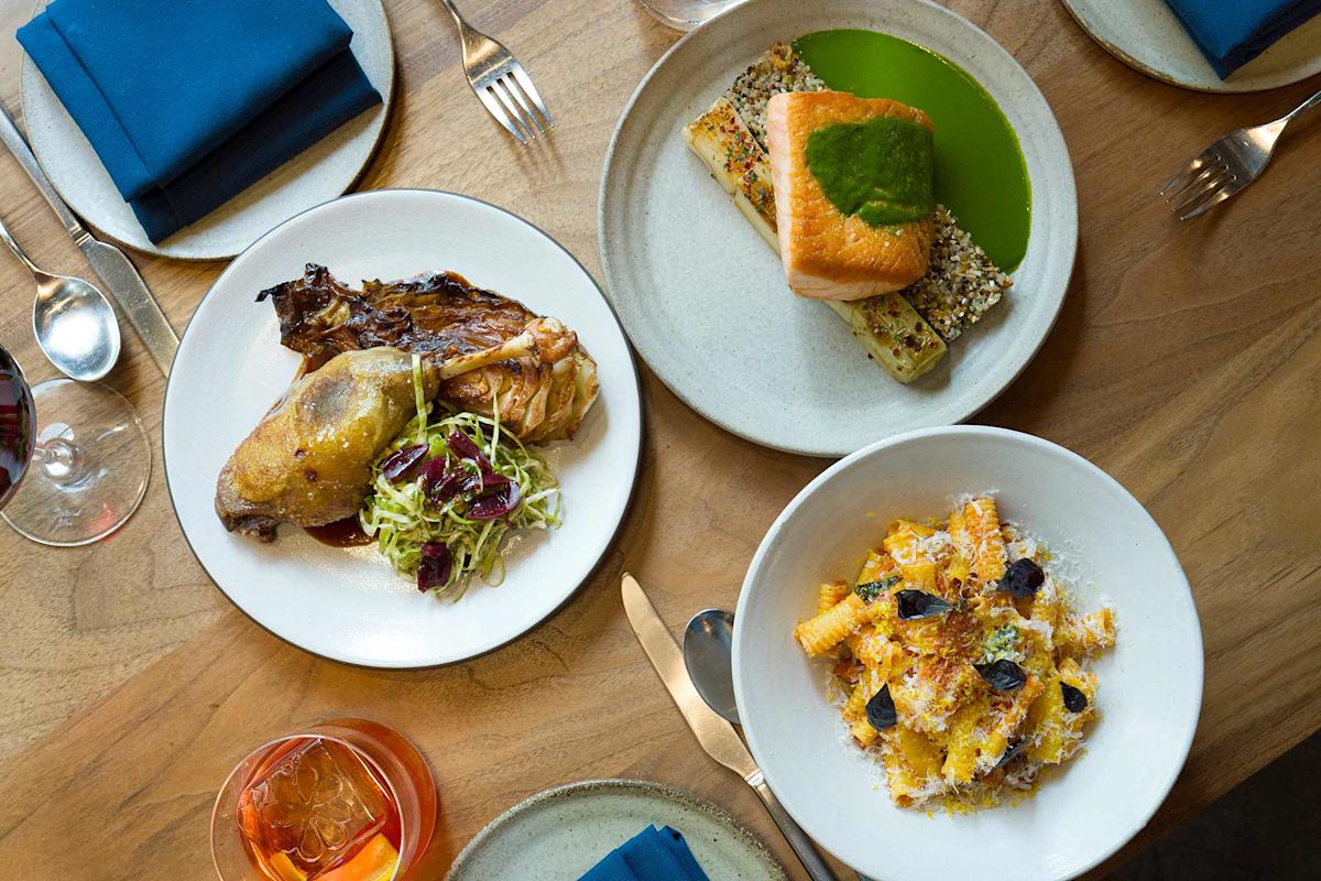 Three plates of gourmet food on a wooden table: roast chicken with greens, baked salmon with grains and green sauce, and a bowl of pasta with cheese. Drinks, cutlery, and blue napkins are also visible.