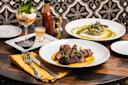 A table set with a plated steak dish, a creamy risotto with asparagus, a dessert topped with mint, a shot glass, and a bottle of hot sauce, against a patterned tile background.