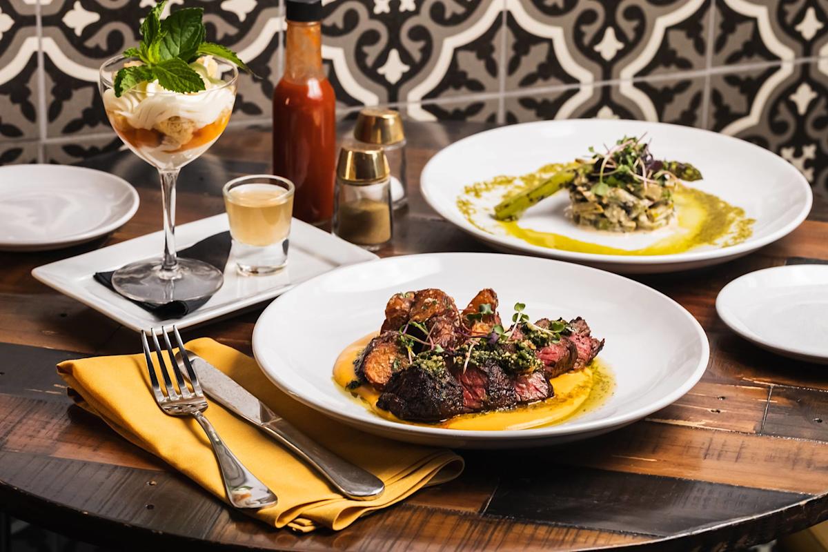 A table set with a plated steak dish, a creamy risotto with asparagus, a dessert topped with mint, a shot glass, and a bottle of hot sauce, against a patterned tile background.