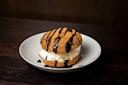 A cream puff filled with vanilla ice cream, drizzled with chocolate sauce, sits on a white plate against a dark wooden background.