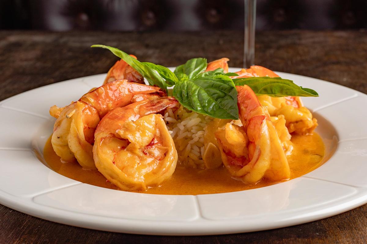 A white plate with shrimp in an orange sauce, served around a mound of white rice and garnished with fresh basil leaves, set on a wooden table.
