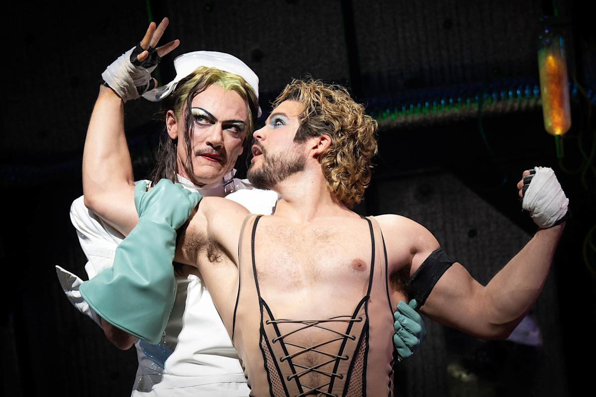 Two actors in dramatic makeup perform on stage. One wears a white costume with gloves, while the other is shirtless with a lace-up corset and dramatic eye makeup. Their expressions are intense and theatrical.